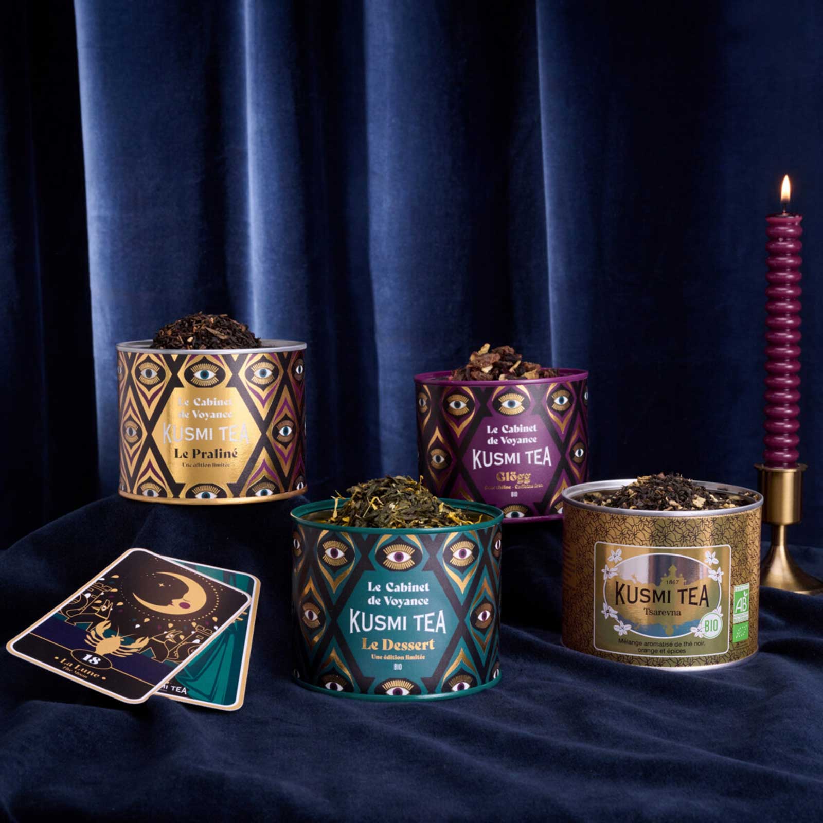 Quatre boîtes de thés Kusmi Tea de la collection Le Cabinet de Voyance disposées devant un rideau bleu sombre avec une bougie et une carte divinatoire