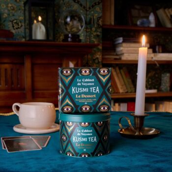 Boîte métallique et coffret de Kusmi Tea Le Cabinet de voyance posés sur une nappe bleue près d'une tasse, d'une bougie allumée et de cartes