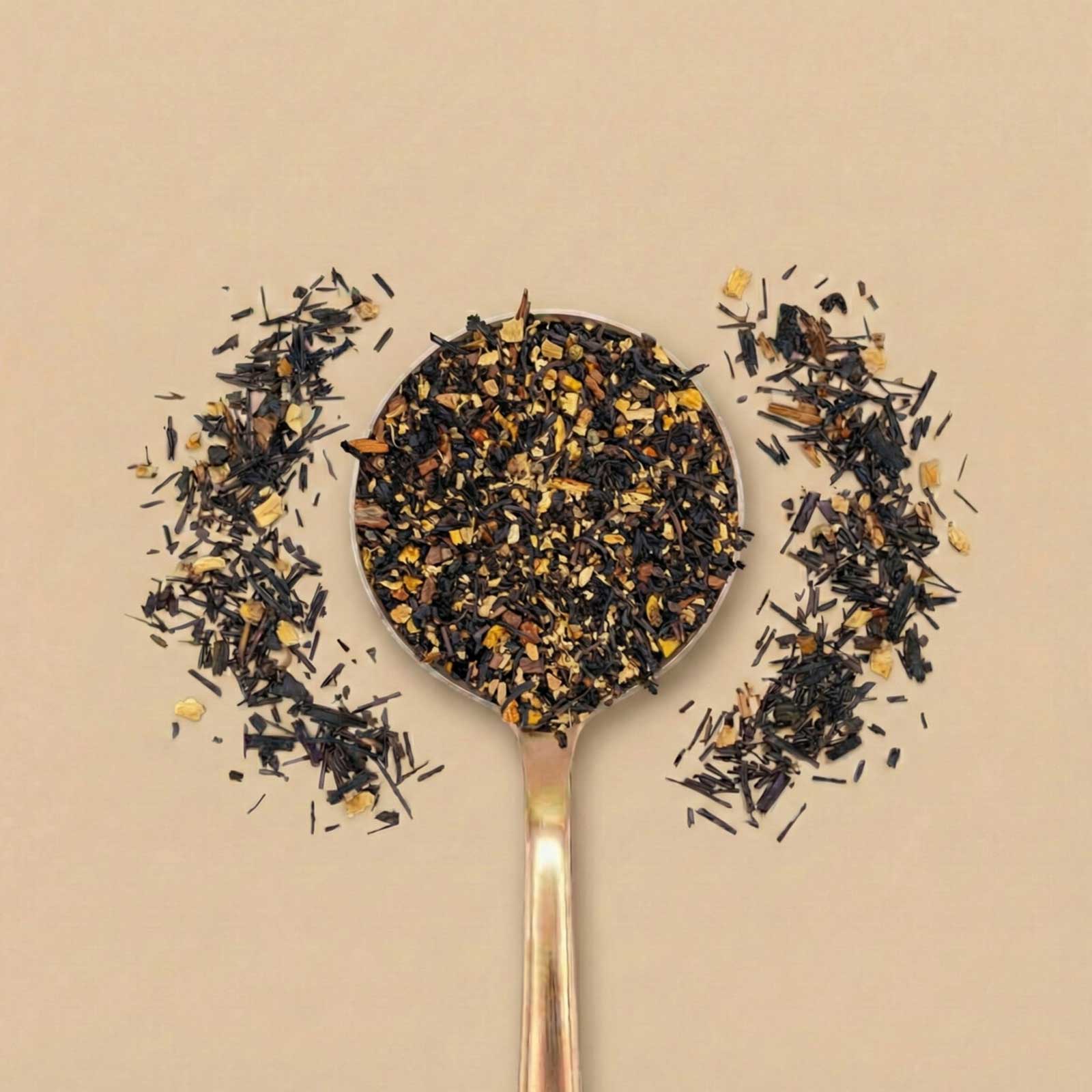 Gros plan d'une cuillère remplie d'un mélange de thé noir et d'épices séchées, entourée de quelques feuilles en vrac sur un fond beige