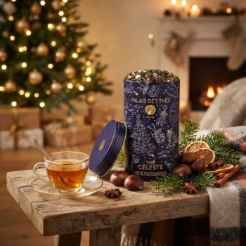 Boîte de thé Thé Céleste de Palais des Thés ouverte sur une table en bois avec une tasse fumante, des châtaignes, des épices et un sapin de Noël